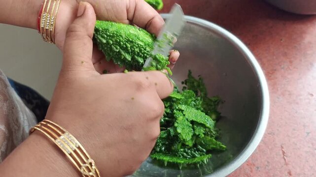 Hands of a person peeling and scraping bitter gourd skin into a stainless steel bowl with a knife close up shot