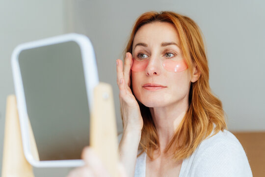Self Cosmetology, skin care, face treatment, spa, natural beauty concept. Smiling middle aged woman in pajamas sits on bed with mirror applying pink moisturizing patches on eyes. Home Beauty routine.