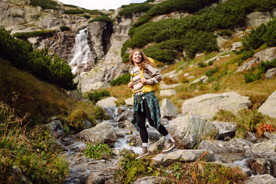 A female hiker with a backpack stands on a path near a waterfall in the mountains. The woman is enjoying nature and the mountain views at sunset. Concepts of nature, hiking, and adventure.