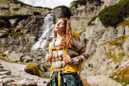 A female hiker with a backpack stands on a path near a waterfall in the mountains. The woman is enjoying nature and the mountain views at sunset. Concepts of nature, hiking, and adventure.