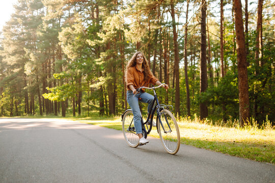 A smiling woman stands on a path in a park with a bicycle. A beautiful woman enjoys a bike ride in a sunny forest. Concepts for a walk, weekend, and relaxation. Active lifestyle.