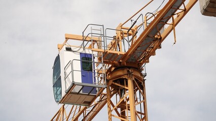 Zbliżenie na biało-niebieską kabinę operatora żurawia wieżowego zamontowaną na żółtej konstrukcji kratownicowej na tle zachmurzonego nieba. © tomo81