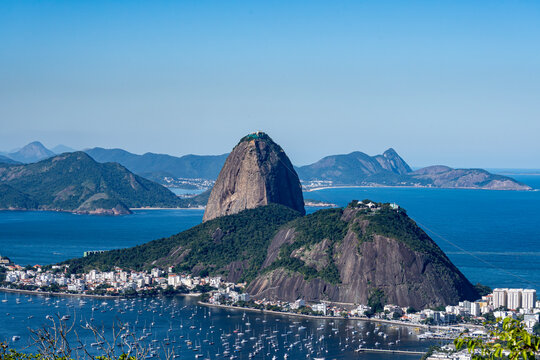 The sweeping view of Sugarloaf Mountain highlights its imposing shape rising above Guanabara Bay, featuring green slopes, blue waters, and the sprawling city. Rio de Janeiro, Brazil.