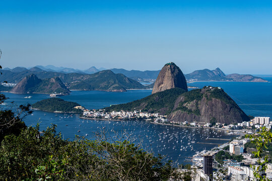 The sweeping view of Sugarloaf Mountain highlights its imposing shape rising above Guanabara Bay, featuring green slopes, blue waters, and the sprawling city. Rio de Janeiro, Brazil.