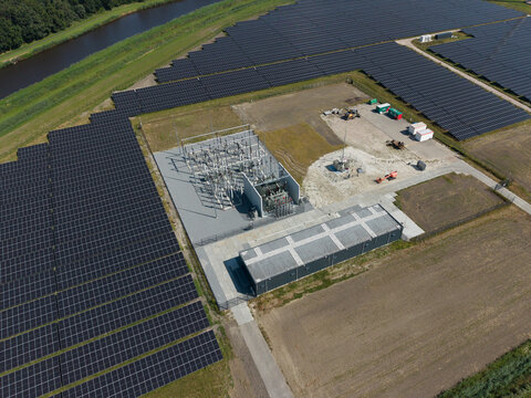 Aerial view of a large solar farm featuring rows of photovoltaic panels and an electrical substation with transformers and switchgear situated next to a canal in the Netherlands.