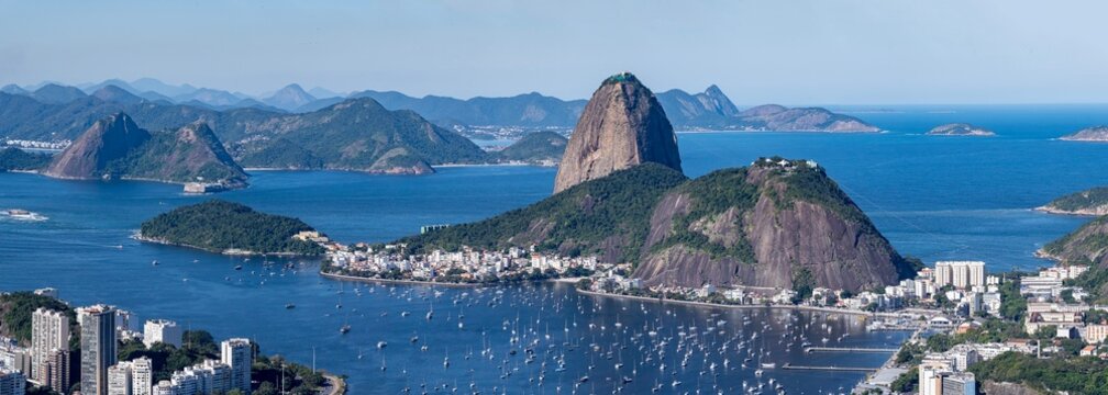 The sweeping view of Sugarloaf Mountain highlights its imposing shape rising above Guanabara Bay, featuring green slopes, blue waters, and the sprawling city. Rio de Janeiro, Brazil.