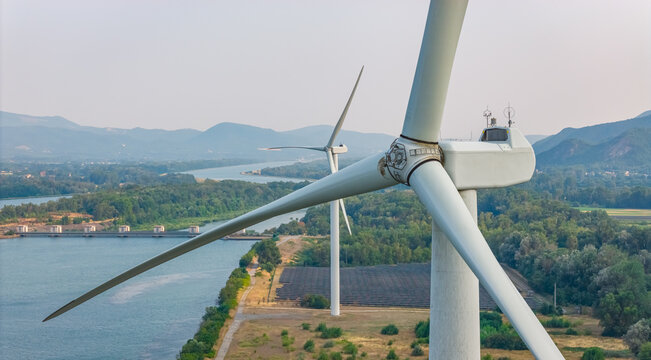 Aerial view of a wind turbine in the foreground with solar panels, the Rhone River dam, a second wind turbine, and a mountain landscape in Le Pouzin, Auvergne-Rhone-Alpes, France.