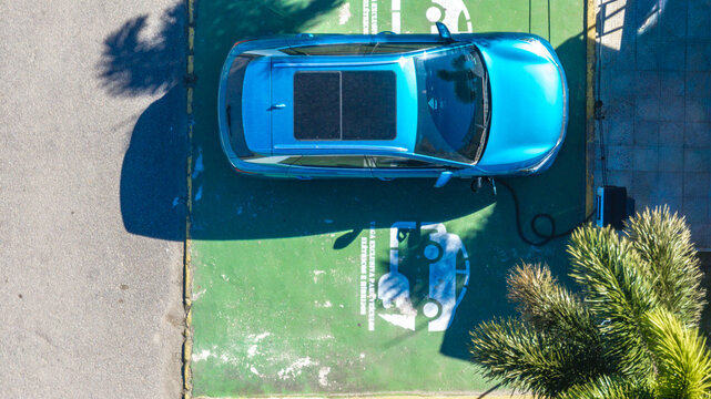 Aerial view of a blue electric car parked in a designated green charging space with a charging cable connected and a palm tree nearby in Brazil.