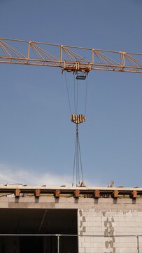 Żuraw budowlany unosi ładunek nad powstającym budynkiem z bloczk&oacute;w na tle bezchmurnego nieba. Widoczny jest mechanizm haka i wysięgnik.