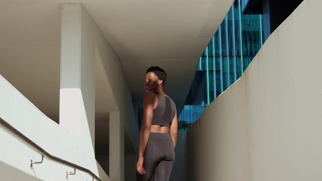Young Black African American woman walks toward her outdoor workout location near modern architecture before beginning fitness training.
