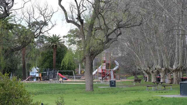 Mature deciduous trees with bare winter branches, a children's playground and park amenities such as picnic tables, benches in Coburg Lake Reserve in Melbourne, Australia