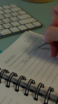 Tracking shot of hand writing in spiral bound journal on desk near computer keyboard in home office close up morning scene