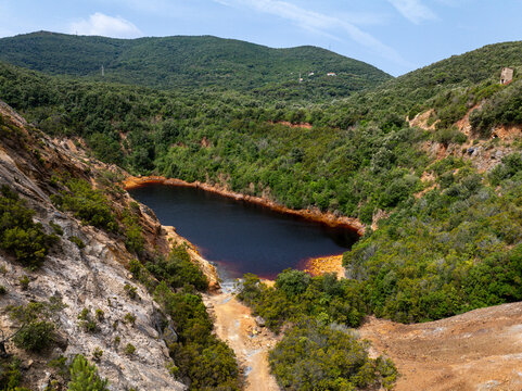 Aerial view of Laghetto delle Conche, also known as Lago di Sangue, featuring its distinctive dark red water surrounded by lush green hills and rocky terrain in Rio Marina, Toscana, Italy.