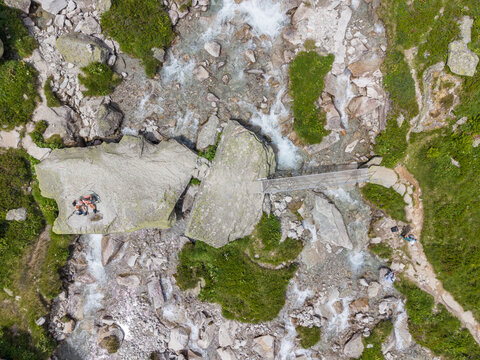 Aerial view of an alpine creek flowing under a mountain footbridge surrounded by rocky terrain and a hiking trail in the mountains of Switzerland.