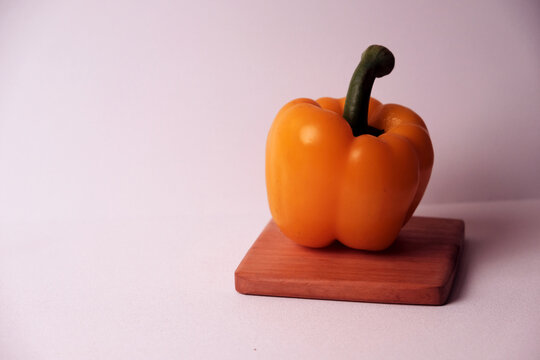 Bright yellow pepper rests on wooden board. Smooth surface gleams under clean white light. Green stem stands tall, contrasting vivid hue. Simple composition draws focus to fresh produce