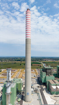 Aerial view of a modern combined cycle natural gas power station with a tall striped chimney stack, industrial buildings, and green fields in Tavazzano con Villavesco, Lombardy, Italy.