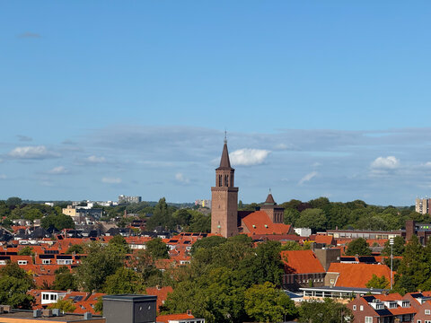 Aerial view of the historic St. Boniface Church tower rising above the red-tiled rooftops and lush green trees of the city under a clear blue sky in Leeuwarden, Friesland, Netherlands.