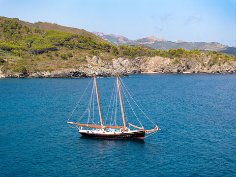 Aerial view of a classic two-masted sailboat cruising on the deep blue waters of the Mediterranean Sea near the rocky coastline and green hills of Lacona, Tuscany, Italy.