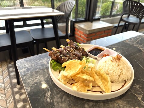 A plate of delicious Saikoro beef satay is served on the table, accompanied by rice, melinjo crackers, and flavorful soy sauce and chili sauce.