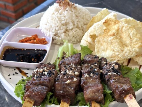 A close-up shot of a delicious plate of Saikoro beef satay served on a table, accompanied by rice, *emping melinjo* (gnetum gnemon chips), and flavorful soy sauce and chili sauce.