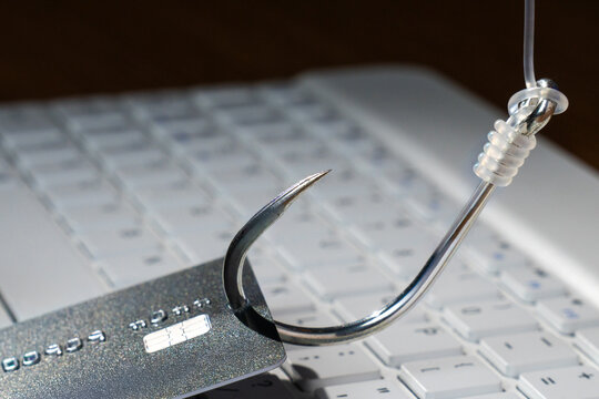 Fishing hook piercing a credit card over a blurred laptop keyboard. Online phishing scams, cybersecurity threats, digital fraud, and internet identity theft concept.