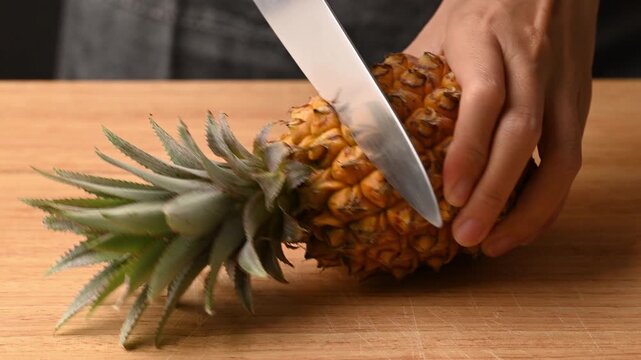 Close up of woman hands using knife to cut top and bottom off fresh pineapple on wooden cutting board