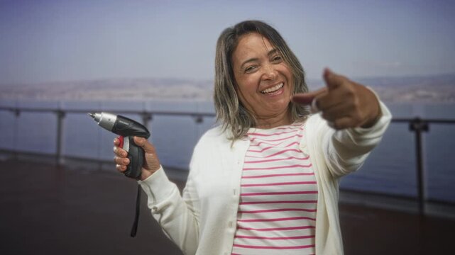 Middle aged woman holding screwdriver and pointing finger while smiling on a building balcony by the water; playful confidence.
