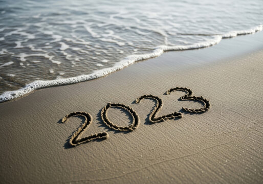 Year number 2023 written in sand on beach shore during sunny day while soft ocean waves wash over text symbolizing passage of time and new start.