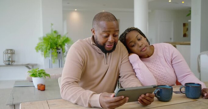 African couple sitting at table, man tapping tablet while woman showing card, confirming payment