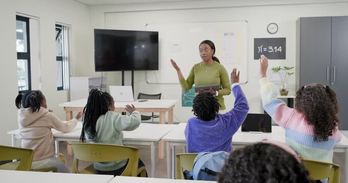African American woman teacher asking in green top with tablet, youth girls raising hands in class