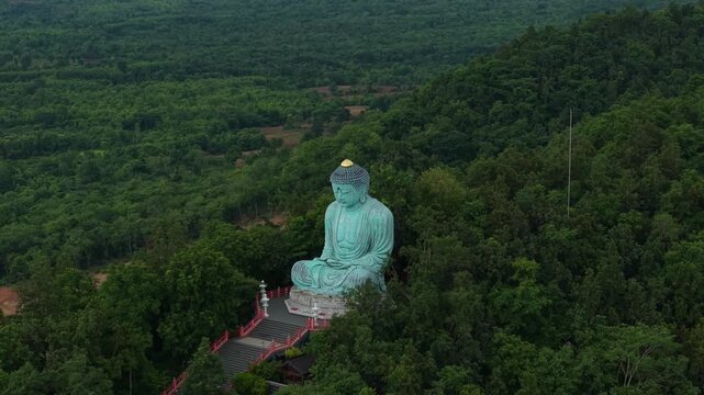 Aerial view of large Daibutsu Buddha statue sitting in meditation surrounded by lush green mountain forest in Lampang, Thailand.