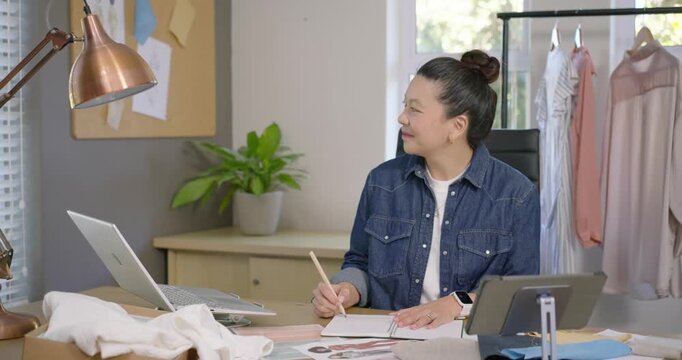 Asian woman checking corkboard, sketching in notebook and referencing tablet in home studio