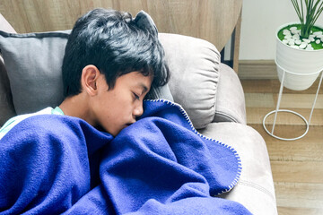 Asian Boy Sleeping Peacefully on Grey Couch, Wrapped in Cozy Blue Fleece Blanket © Gatot