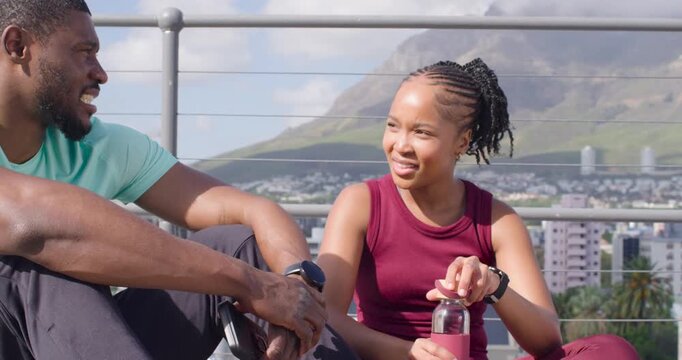 Workout partners sitting rooftop, woman fidgeting cap, opening pink bottle, man reaching to drink
