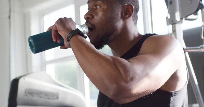 Mid 30s African man ending set opening teal bottle showing yellow tab drinking recovering at gym