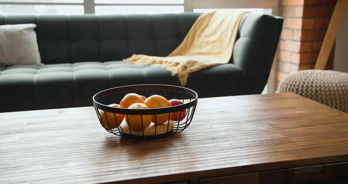 Camera is pulling back, revealing wire basket of oranges and apple on wood table, establishing calm
