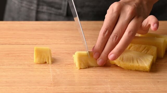 Close up of woman hands using a knife to slice fresh pineapple into small pieces on a wooden cutting board