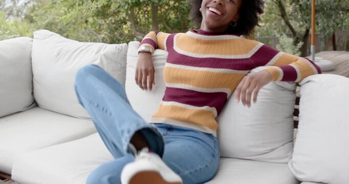 Woman stepping into frame on patio, lowering onto sofa in striped sweater adjusting pose for camera