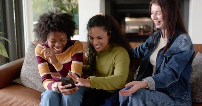 Female friends on couch with left woman holding smartphone, leaning in and laughing at content