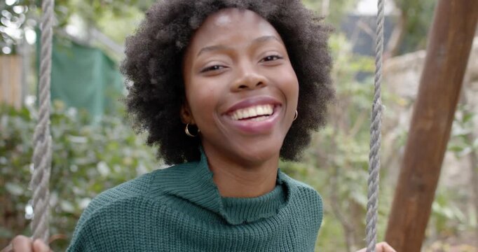 African woman in garden holding rope swing, laughing as camera moving closer, wearing green sweater