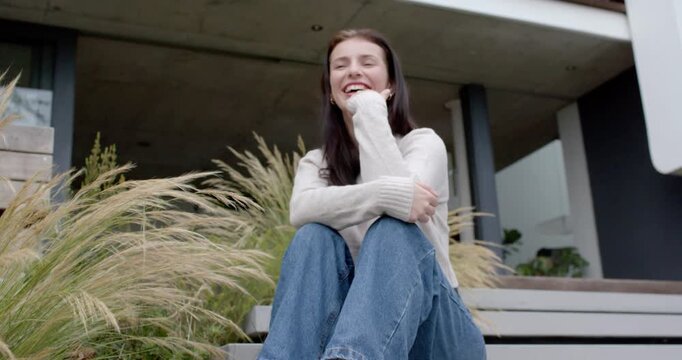 Camera revealing woman sitting on front steps, hugging knees, touching chin, smiling then laughing