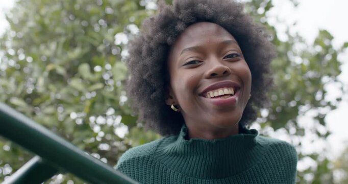 Seeing camera, African American woman leaning on green railing at park, smiling, posing in sweater