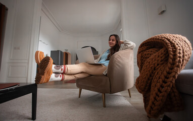 Young female freelancer reclining in an armchair with her feet raised on a table and laptop on her...