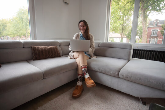 Smiling young woman working remotely on laptop from home sofa