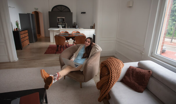Young woman looking at camera while reclining in an armchair with laptop and feet raised. Wide view of flexible remote work and a comfortable open-plan home office lifestyle.