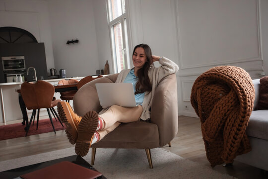 Smiling young woman reclining comfortably in an armchair with one hand behind her head while using a laptop. Flexible remote work and stress-free home office concept.