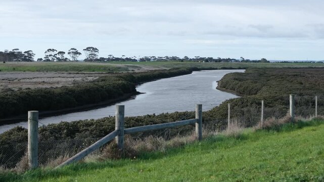 Hovells Creek winding through coastal saltmarsh and open rural terrain in Victoria, Australia. Wooden post and wire fence runs along a grassy hill, calm creek bordered by low coastal vegetation.