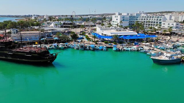 Drone view of Ayia Napa Harbour in Cyprus. Blue marina water, yachts, harbour buildings and coastal roads shape a lively waterfront panorama.