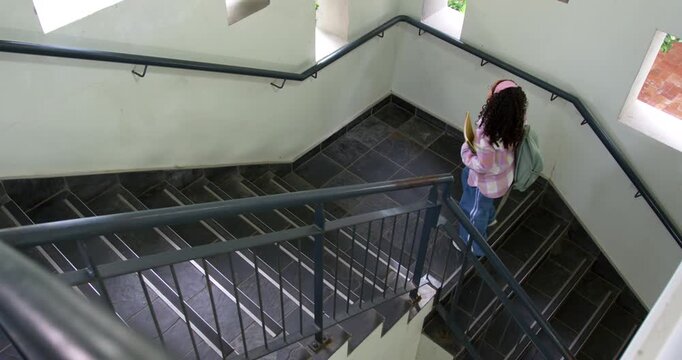 African American teen glancing at notes descending stairs, going to class with backpack, copy space