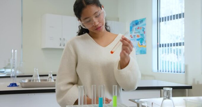 Child in goggles spotting red droplet, examining test tube, adding liquid using pipet at school lab